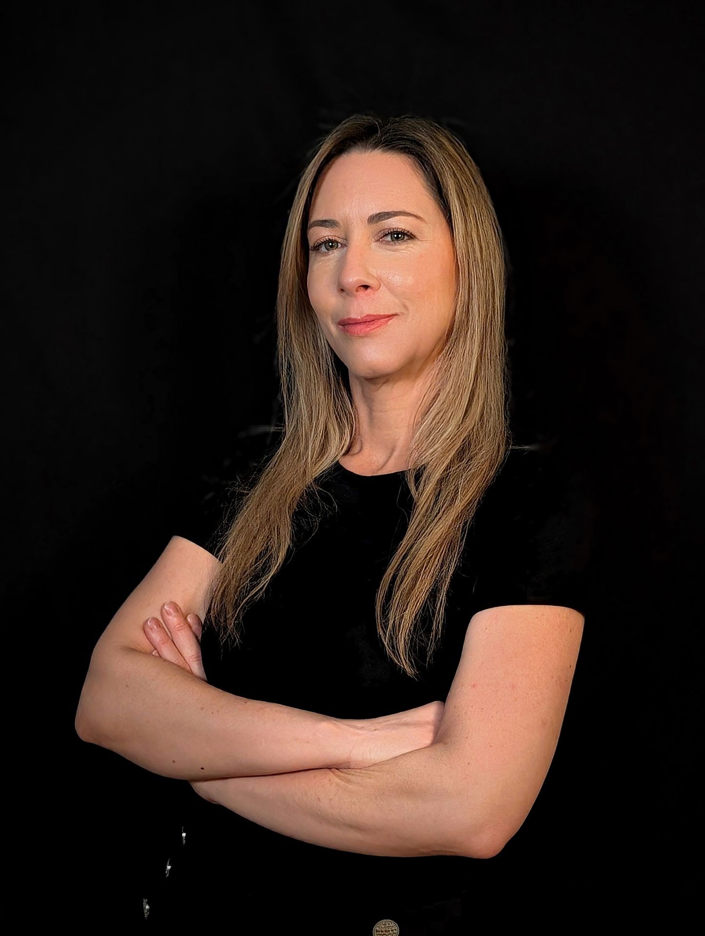 Woman with long hair in black shirt crossing arms against a dark background.