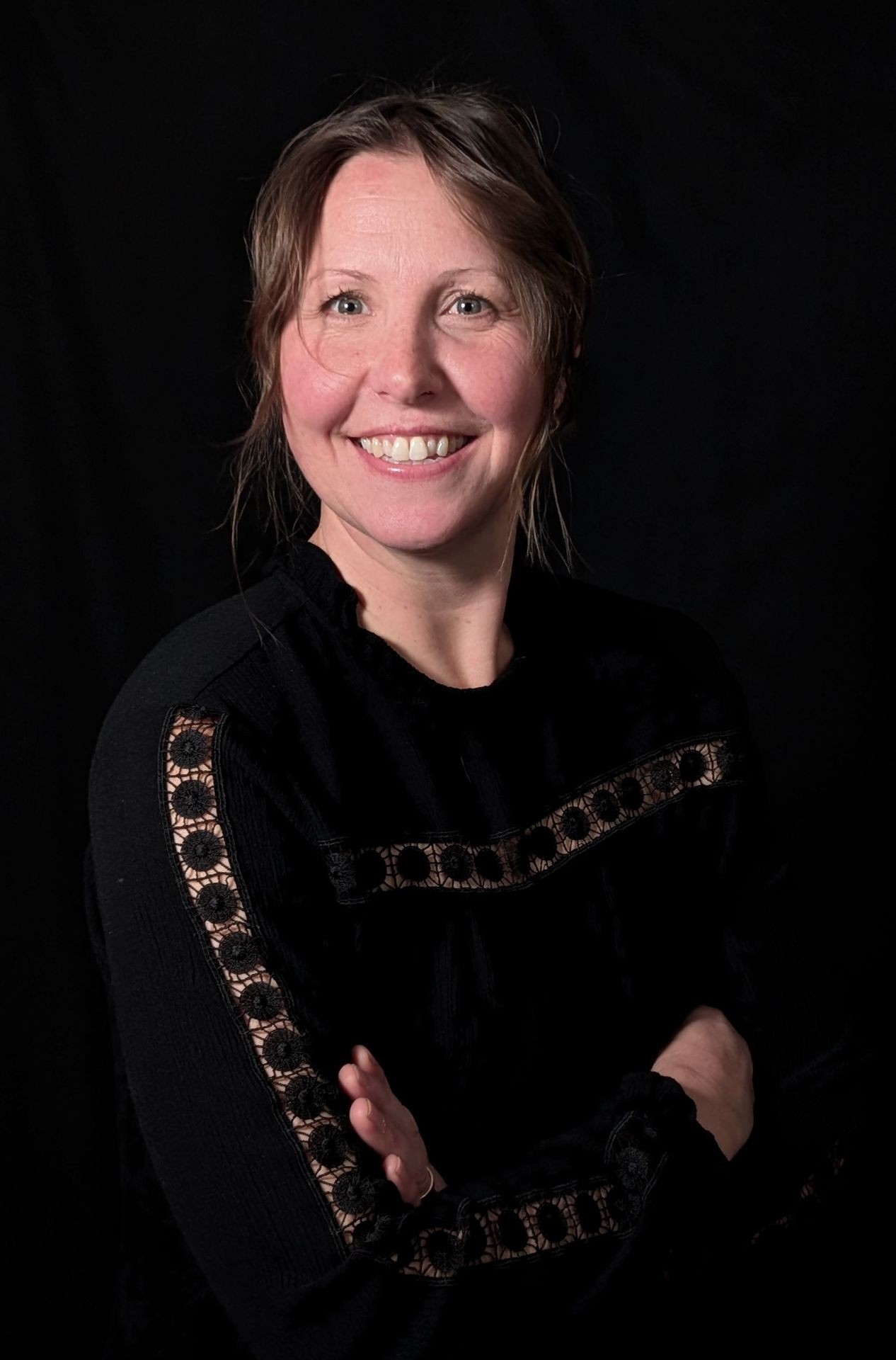 Woman smiling with crossed arms, wearing a black top with lace details, against a dark background.